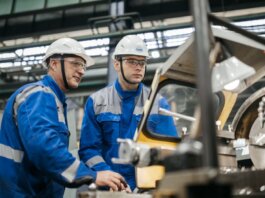 Two industrial workers in protective gear operate machinery in a factory setting.
