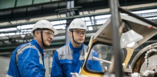 Two industrial workers in protective gear operate machinery in a factory setting.