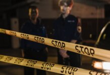 Two police officers standing behind a stop tape at night, scene partially illuminated by car headlights.