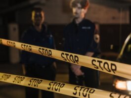 Two police officers standing behind a stop tape at night, scene partially illuminated by car headlights.