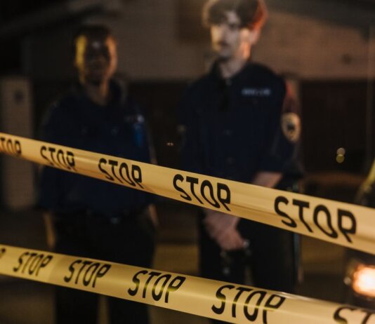 Two police officers standing behind a stop tape at night, scene partially illuminated by car headlights.