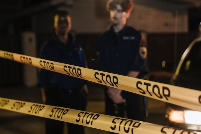 Two police officers standing behind a stop tape at night, scene partially illuminated by car headlights. Two police officers standing behind a stop tape at night, scene partially illuminated by car headlights.