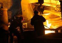 Two workers operate in a steel factory, managing molten metal in an industrial furnace.