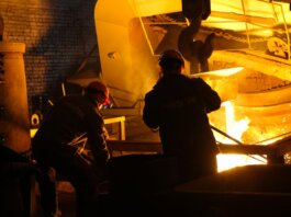 Two workers operate in a steel factory, managing molten metal in an industrial furnace.