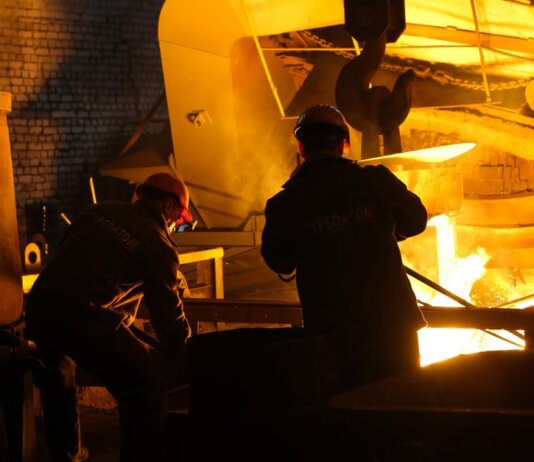 Two workers operate in a steel factory, managing molten metal in an industrial furnace.