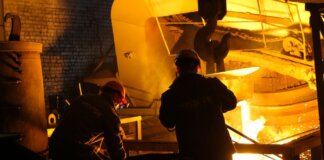 Two workers operate in a steel factory, managing molten metal in an industrial furnace.