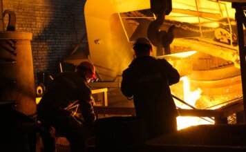 Two workers operate in a steel factory, managing molten metal in an industrial furnace.