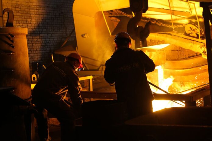 Two workers operate in a steel factory, managing molten metal in an industrial furnace. Two workers operate in a steel factory, managing molten metal in an industrial furnace.