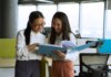 Canada Summer Jobs 2027: работодатели могут подавать заявки до 3 декабря 2026 года Two young women engaged in a collaborative discussion in a bright, modern office setting.