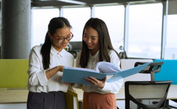 Canada Summer Jobs 2027: работодатели могут подавать заявки до 3 декабря 2026 года Two young women engaged in a collaborative discussion in a bright, modern office setting.