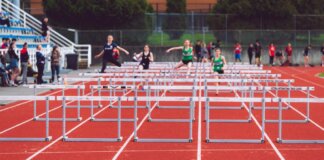 Women athletes compete in a hurdles race at a Victoria track event on a sunny day.