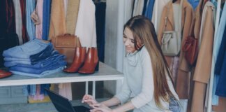 Young woman managing a fashion boutique, multitasking with phone and laptop, surrounded by clothing.